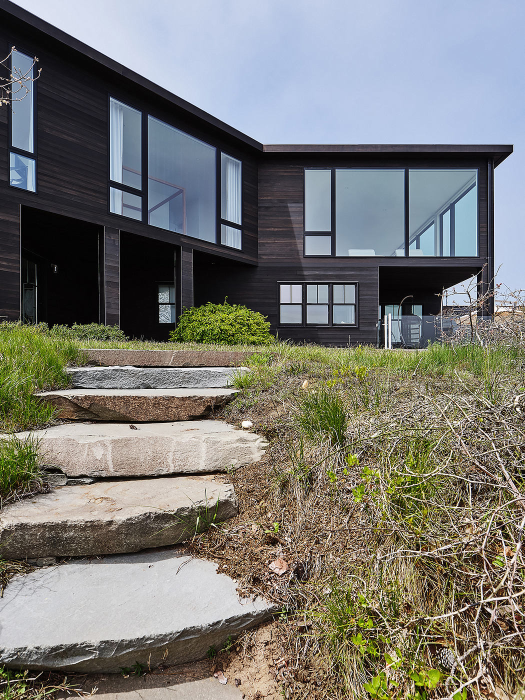 A modern, dark-colored house with large windows, a staircase, and a grassy surrounding.