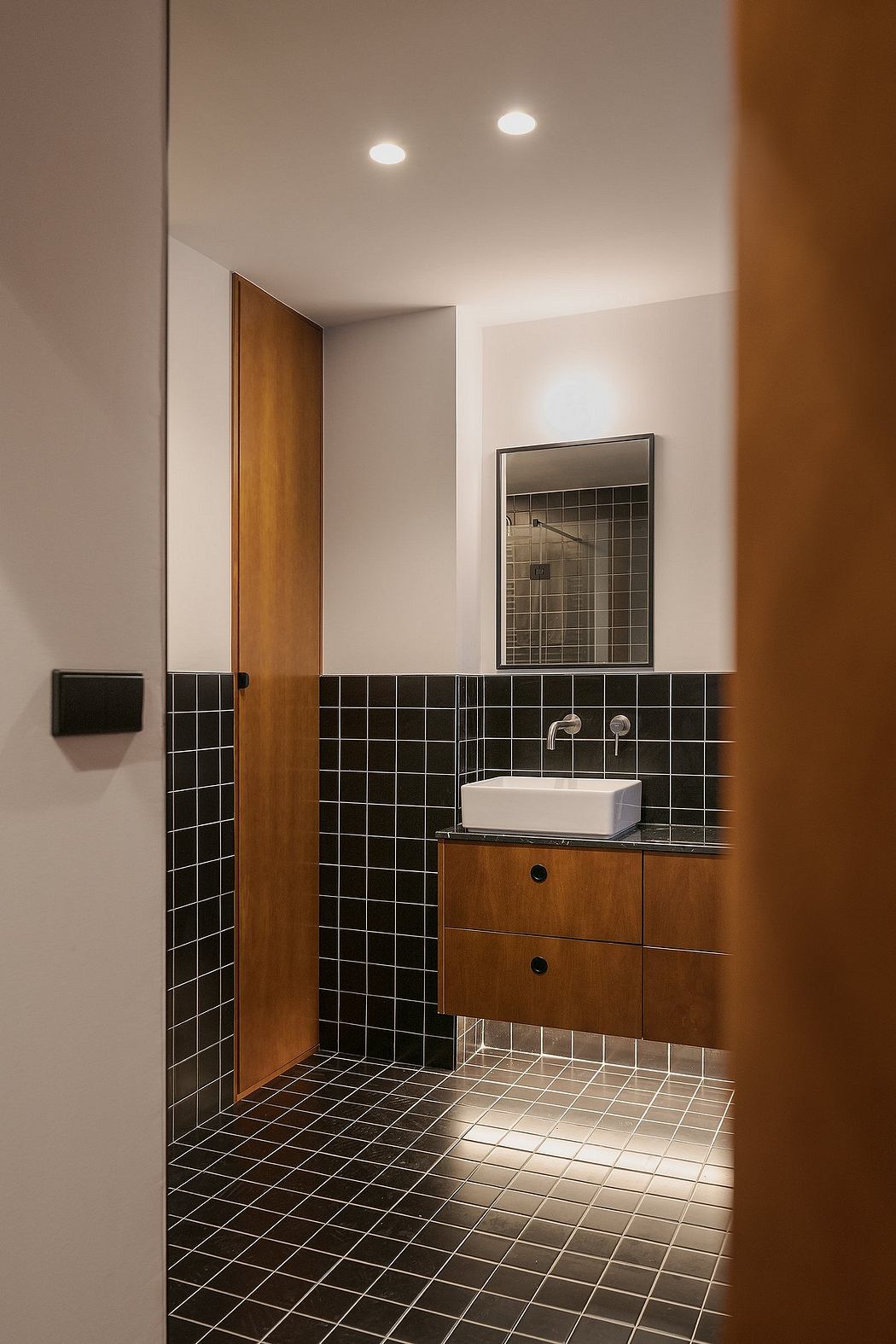 A modern bathroom with wooden vanity, black tile walls, and recessed lighting.