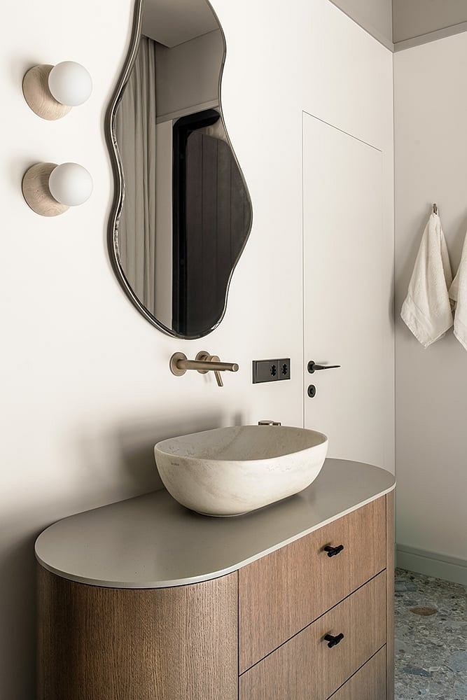 Minimalist bathroom design with curved mirror, light fixture, and wood vanity cabinet.