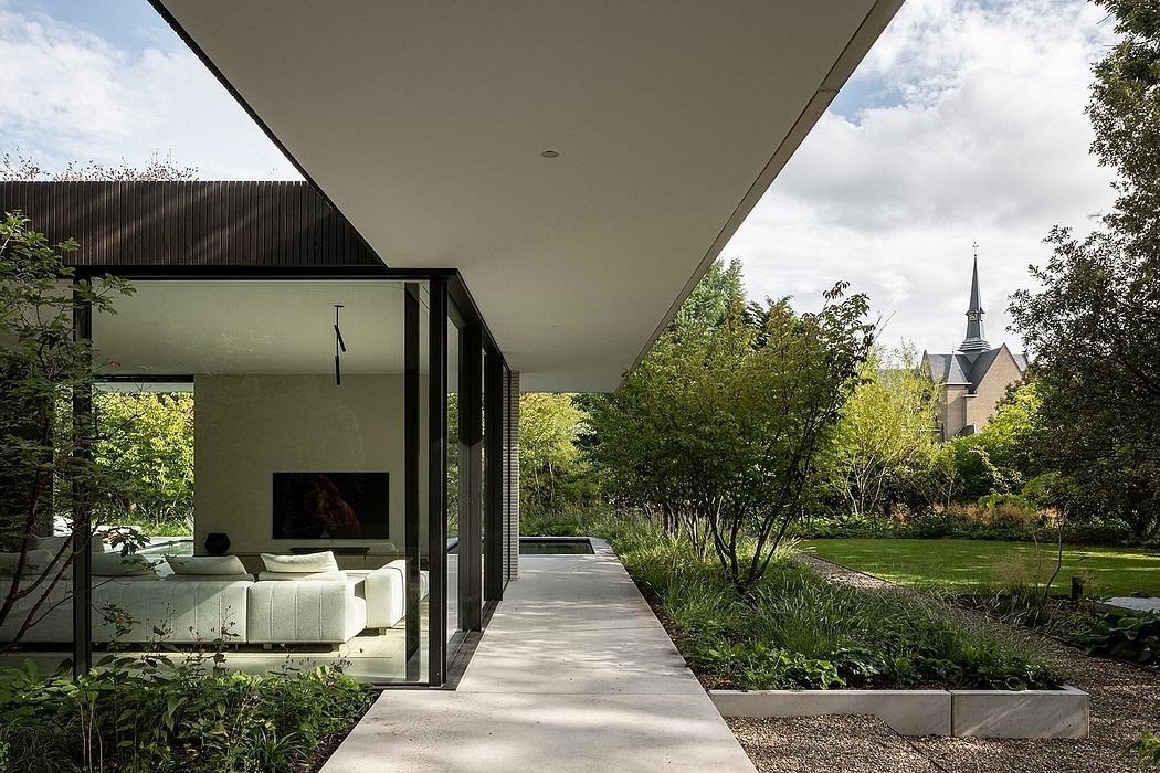 Modern home with glass walls, sleek outdoor walkway, and lush garden with church spire.