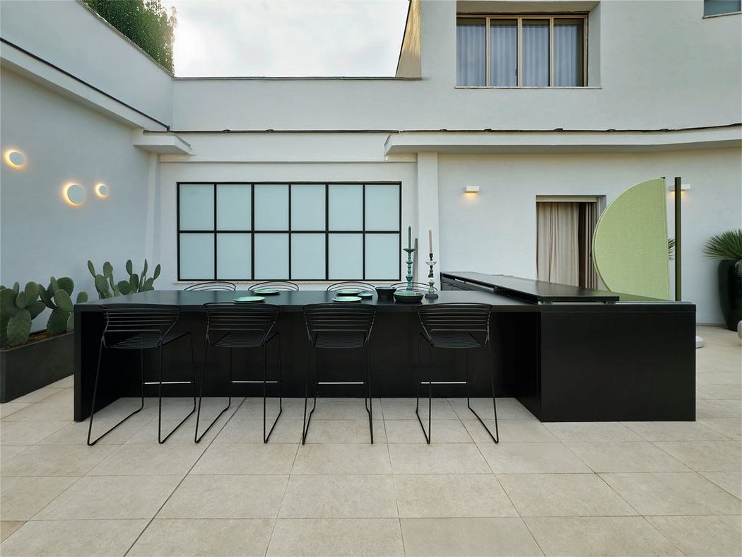 A modern, minimalist outdoor dining area with a sleek black table and chairs, surrounded by large windows and plants.