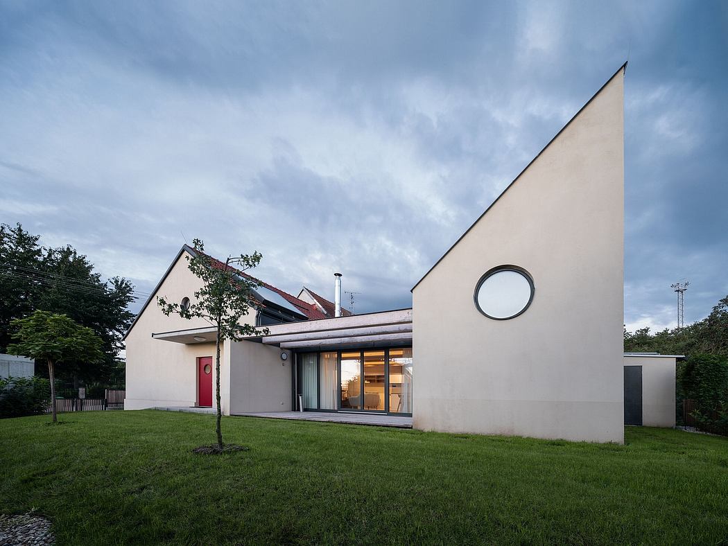 Modern residential building with triangular roof and circular window, surrounded by lush greenery.