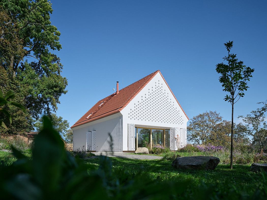 A modern, white house with a red tile roof and perforated exterior walls, surrounded by lush greenery.