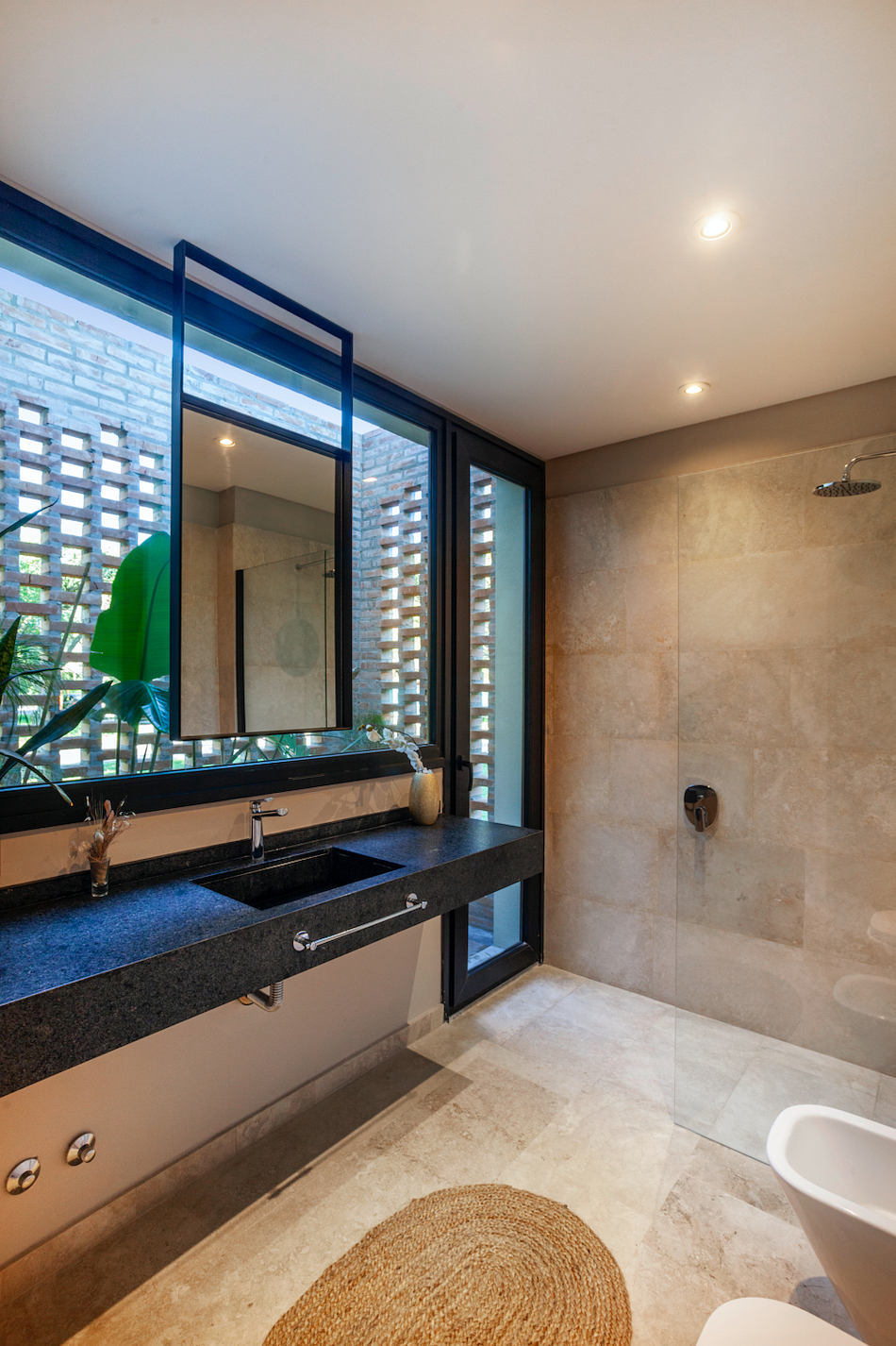 Sleek modern bathroom with black stone vanity, large glass shower, and natural stone tiles.