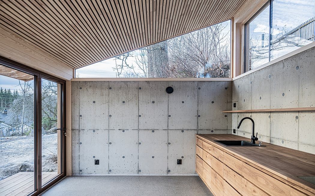 Open-plan interior with concrete walls, wooden cabinetry, and large windows overlooking nature.