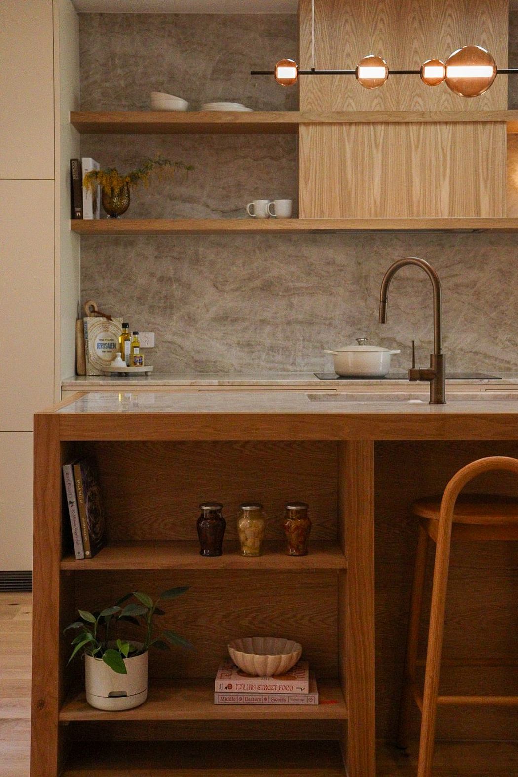 A cozy kitchen with wood shelving, a bronze light fixture, and a neutral stone backsplash.
