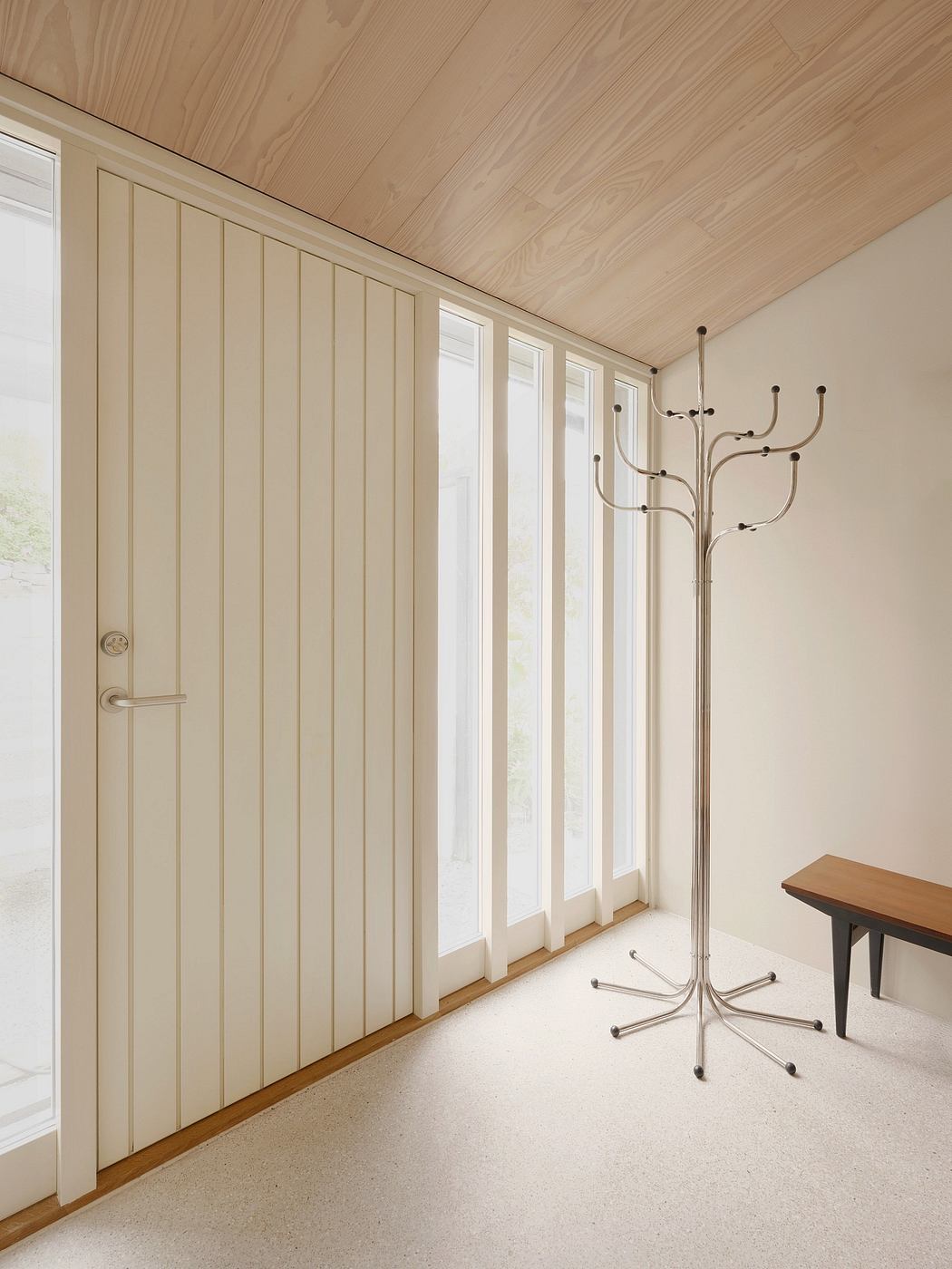 Warm-toned wooden paneled walls, tall windows, and a modern metal coat rack highlight the minimalist design.