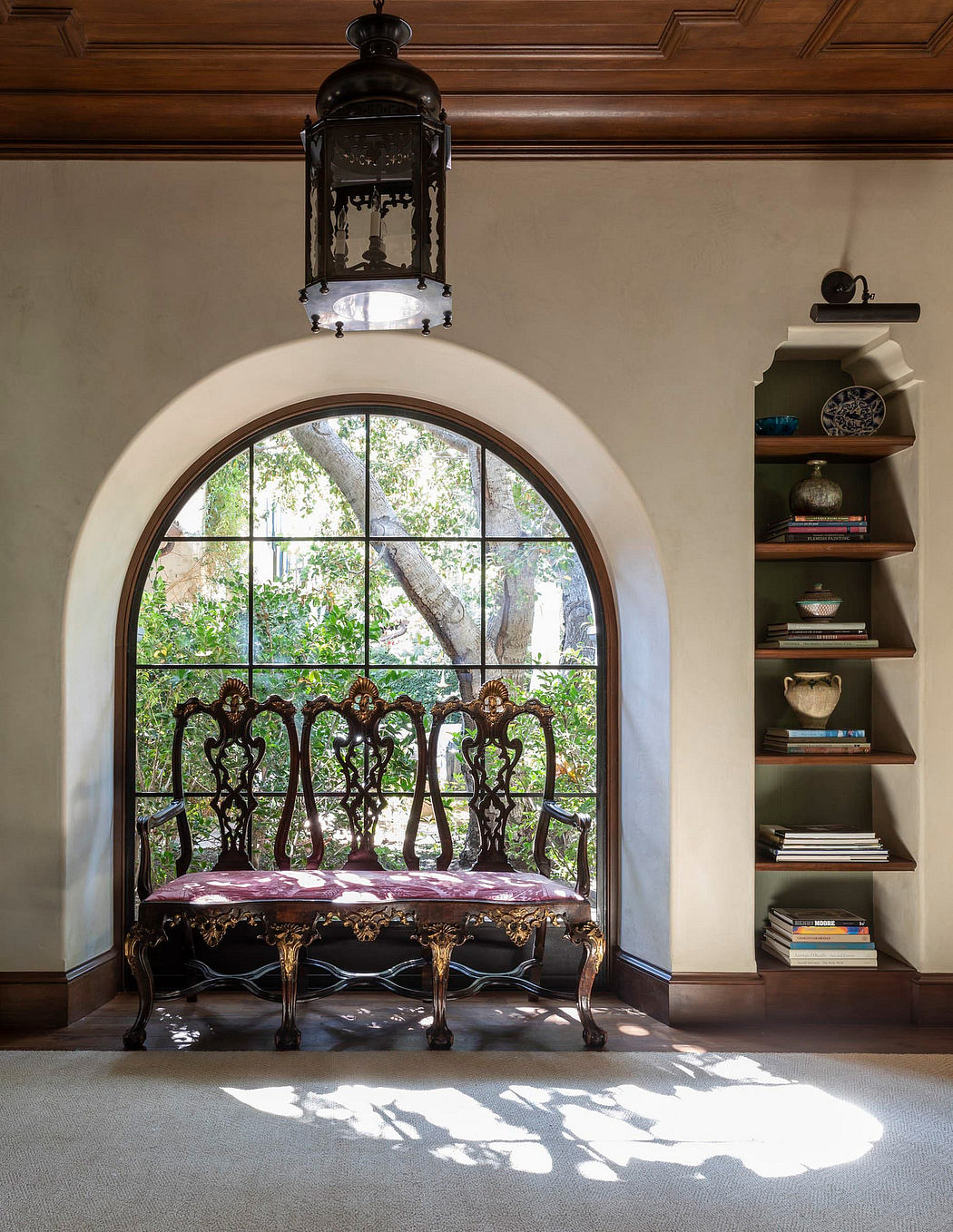 A cozy nook with an ornate wrought-iron bench beneath a grand arched window framing lush greenery.