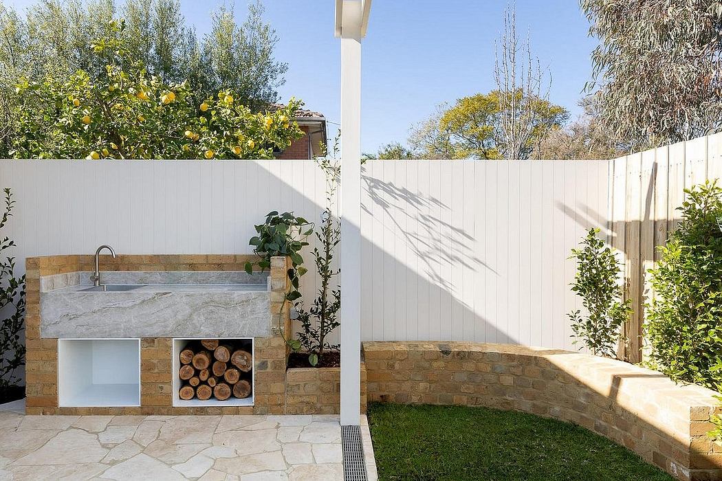 Cozy outdoor kitchen with stone counter, wood storage, and lush greenery surrounding.