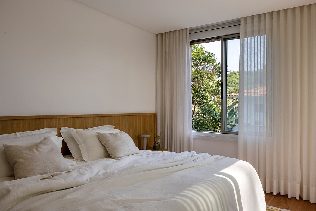 Minimalist bedroom with wooden headboard, large window, and sheer curtains overlooking greenery.