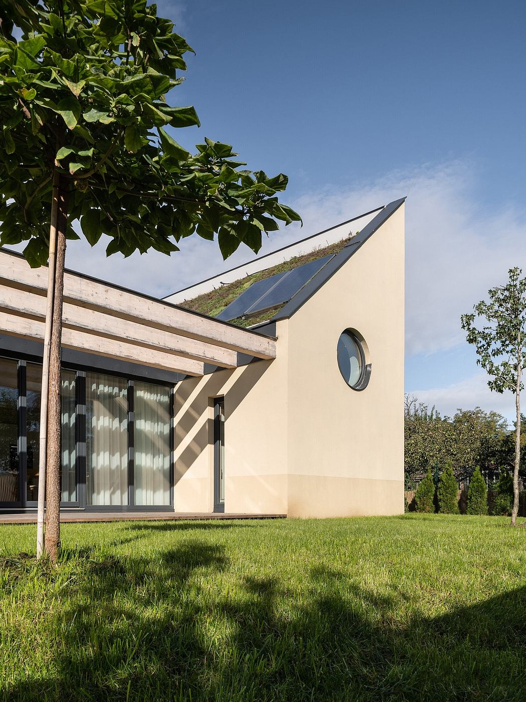A modern, eco-friendly house with a green roof, circular window, and wooden beams.