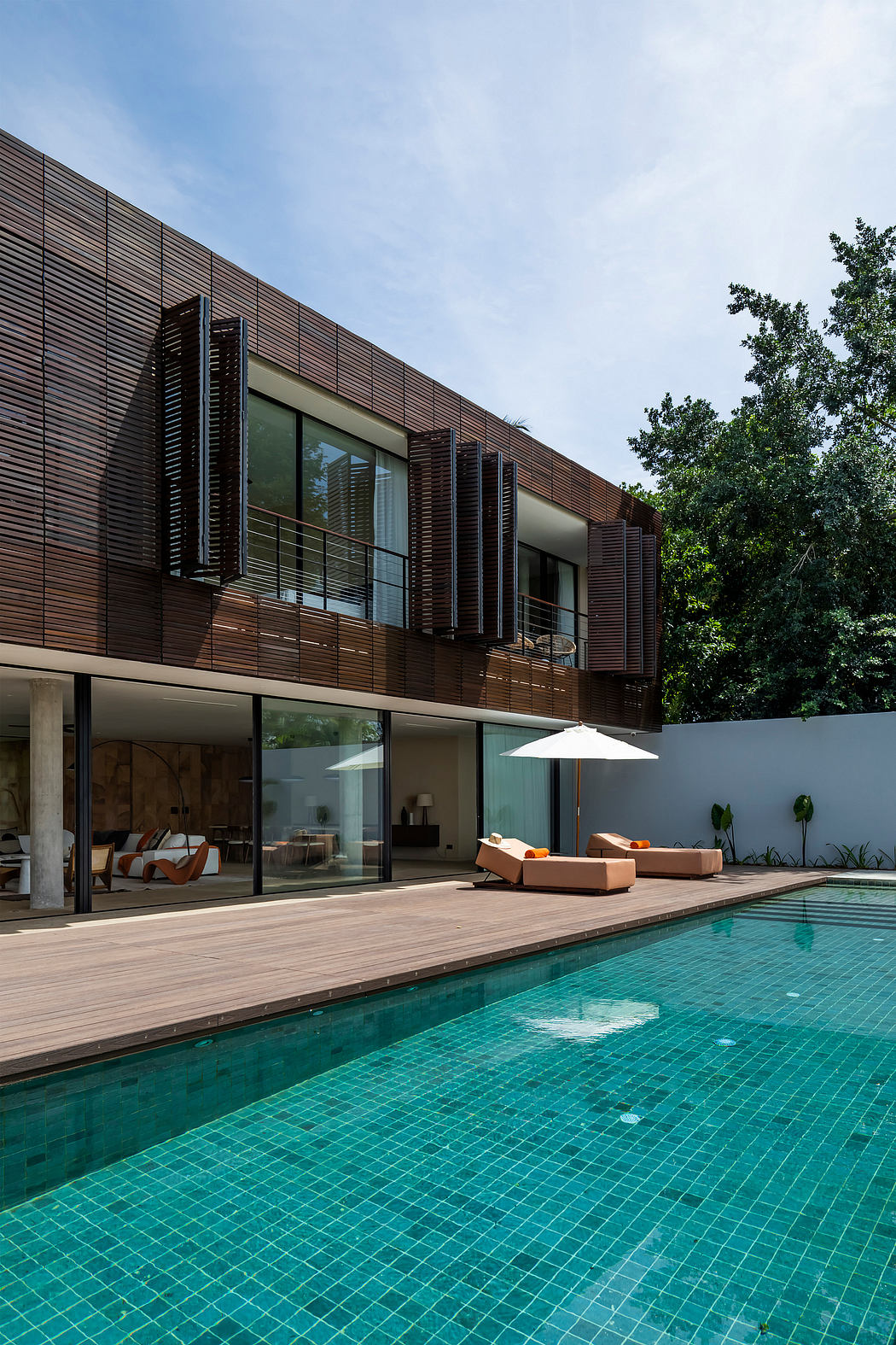 Two-story modern glass and wood house with pool and lounge area surrounded by trees.