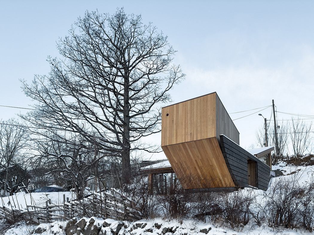 A modern, wood-paneled cabin-like structure with a cantilevered upper level, set against a snowy, wooded backdrop.