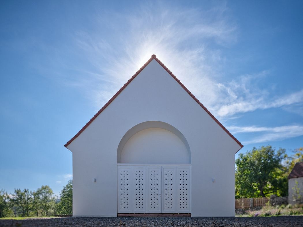A simple, white building with a triangular roof and a semicircular entryway featuring perforated panels.